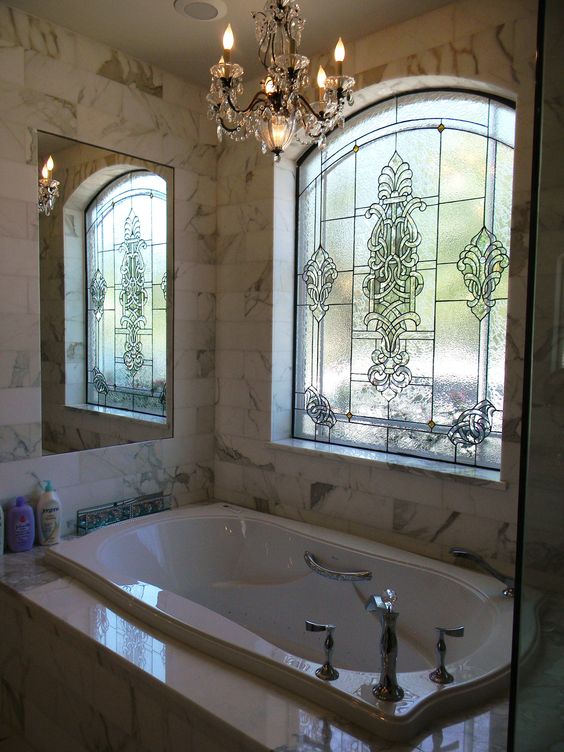 a bathtub clad with marble tiles, a mirror, a crystal chandelier and a stained glass window to keep privacy yet provide light