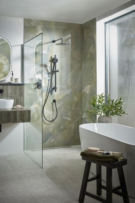 a contemporary bathroom with green onyx tiles, an oval tub, a dark-stained stool and a floating vanity with skinny tiles