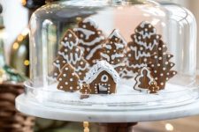 a large cloche with a wooden stand and glazed gingerbread houses and mini Christmas trees is a Christmas terrarium