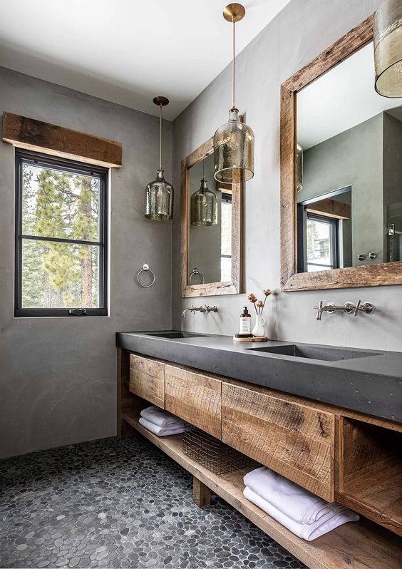 a catchy bathroom with concrete walls and a vanity, wooden storage units, pendant lamps and mirrors in rough wooden frames