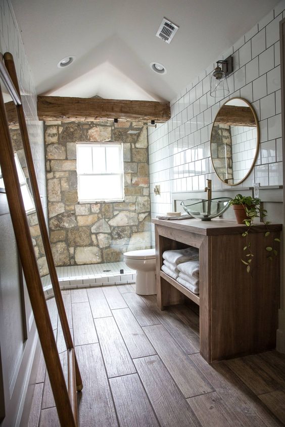 a chalet bathroom with a stone accent wall, a wooden beam, a wooden vanity and a round mirror looks very cozy