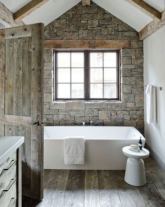 a chalet bathroom with a stone accent wall, wooden beams on the walls and ceiling, a bathtub and a side table plus a window