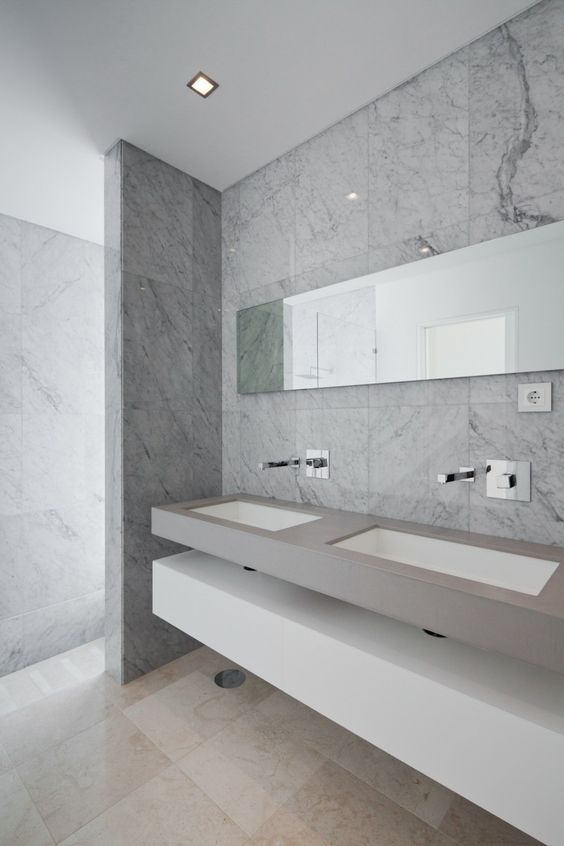 a chic minimalist bathroom with marble patterned tiles, a floating concrete vanity with sinks and a white slab shelf plus a long mirror