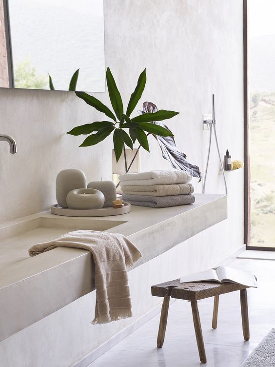 a concrete bathroom with a floating concrete vanity and a sink, a wooden stool, large mirrors and pretty towels
