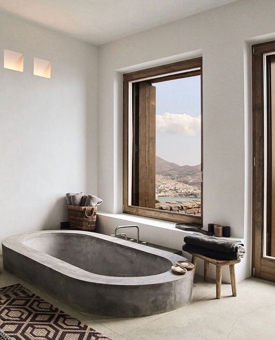 A contemporary bathroom with windows fo a view, a concrete floor and a built in concrete bathtub, a printed rug and wooden sotols