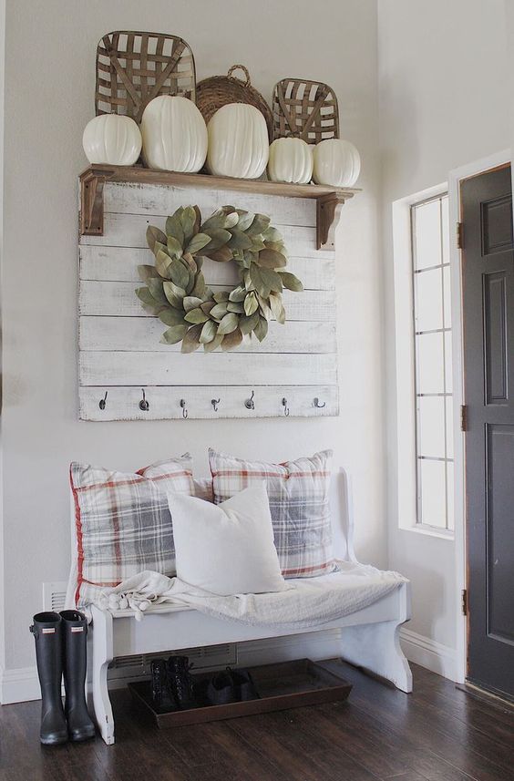 a farmhouse entryway with a white bench, checked pillows, a white rack, a wreath and a rack with white pumpkins for fall