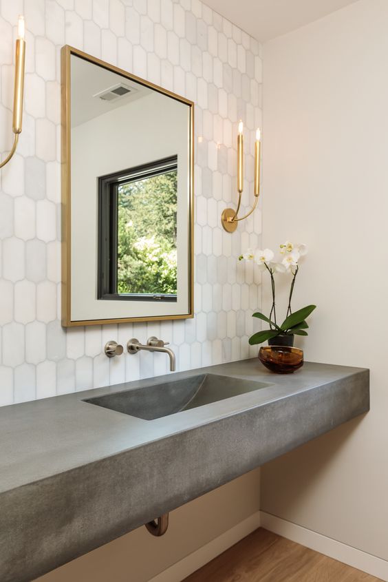 A glam bathroom with a white tile wall, a concrete vanity with a built in sink, a mirror in a gilded frame and gilded sconces