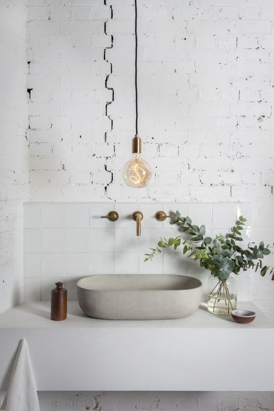 a minimalist white bathroom with a brick wall, a floating vanity with a concrete sink, a bulb and brass fixtures