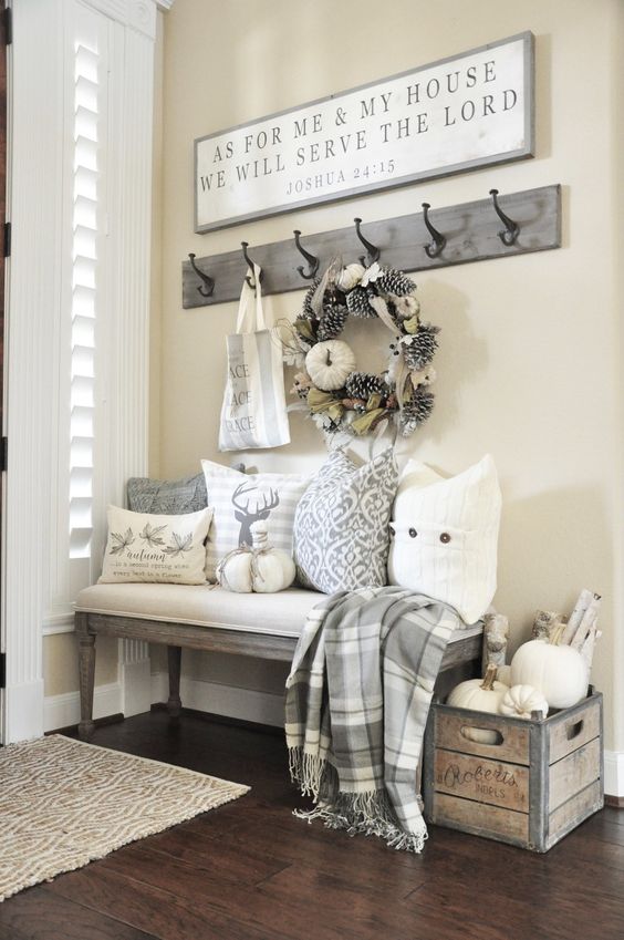 a neutral farmhouse entryway with an upholstered bench, printed pillows, a crate with branches and pumpkins, a rack with hooks and a sign