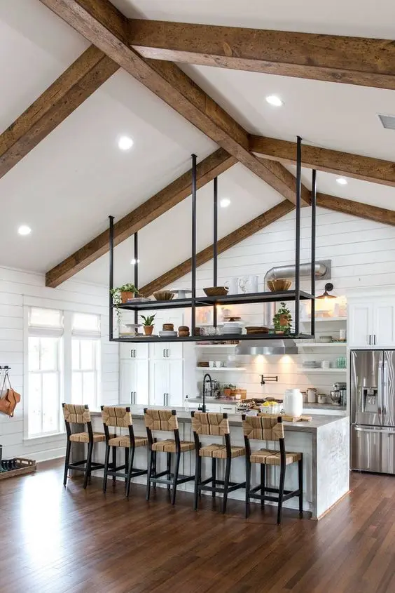 a neutral farmhouse kitchen clad with beadboard, with white cabinetry, woven stools, wooden beams and a pendant storage unit