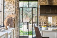 a rustic kitchen with stone walls, wooden beams and ceiling for much coziness and white cabinetry plus leather chairs