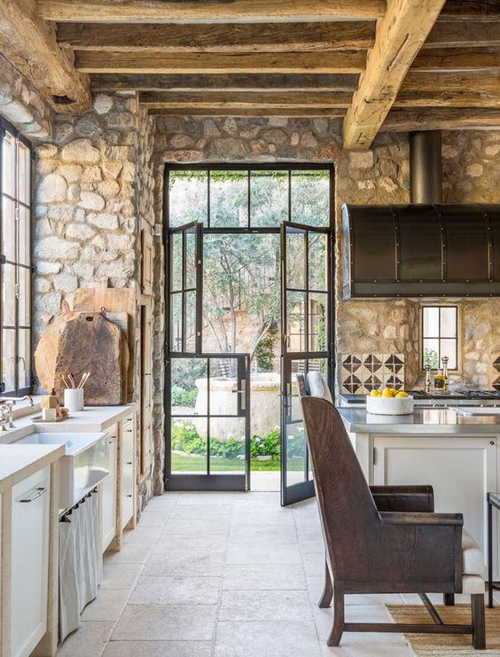 a rustic kitchen with stone walls, wooden beams and ceiling for much coziness and white cabinetry plus leather chairs