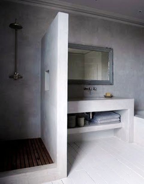 A sleek minimalist concrete bathroom with a built in vanity, a shower space with a wooden floor and a tub clad with concrete