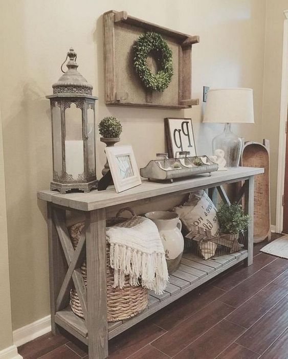 a weathered wood rustic console table with a basket with blankets, a candle lantern, some boxwood and a dough bowl