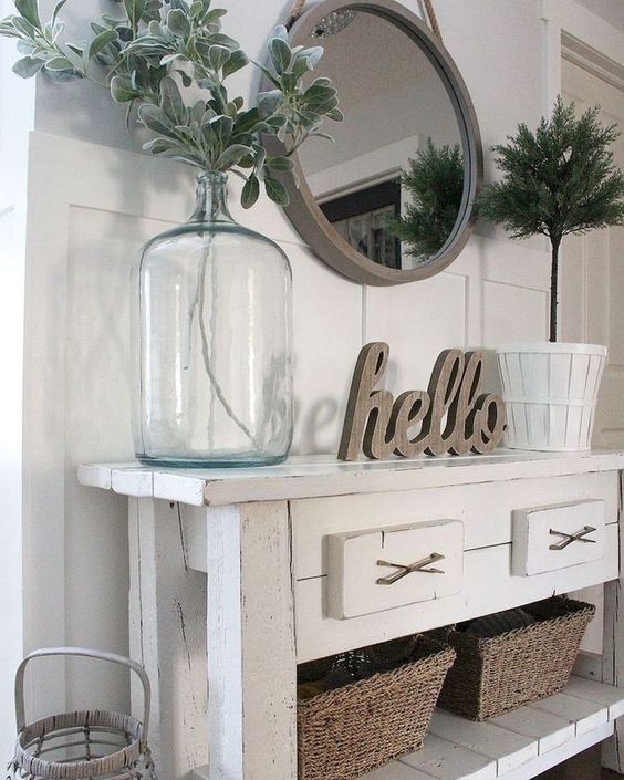 a white console table, baskets for storage, a round mirror in a wooden frame and some greenery in a bottle