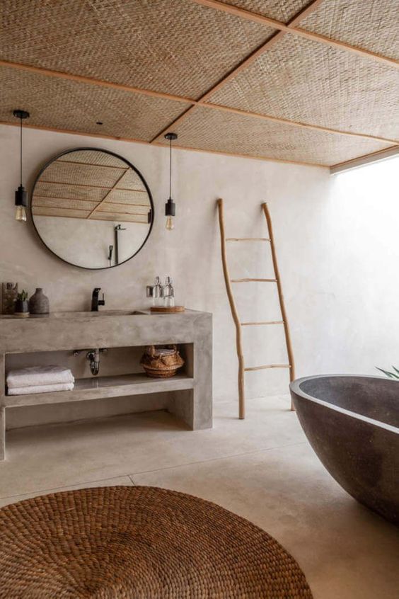 an earthy bathroom clad with concrete, with a stone bathtub, a concrete vanity, a round mirror, bulbs and a rug plus a woven ceiling