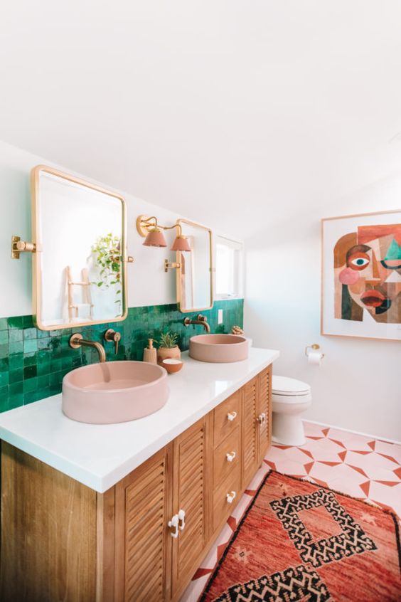 a colorful mid-century modern bathroom with green tiles for a backsplash, mosaic ones on the floor, pink sinks and lamps and a catchy wall art