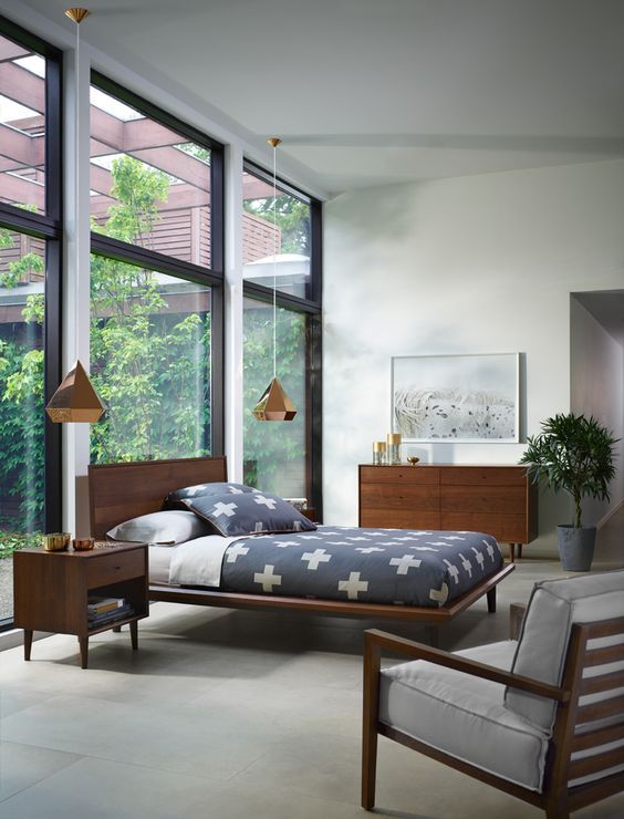 A light filled mid century modern bedroom with a glazed wall, rich stained wooden furniture, pendant lamps and a potted plant