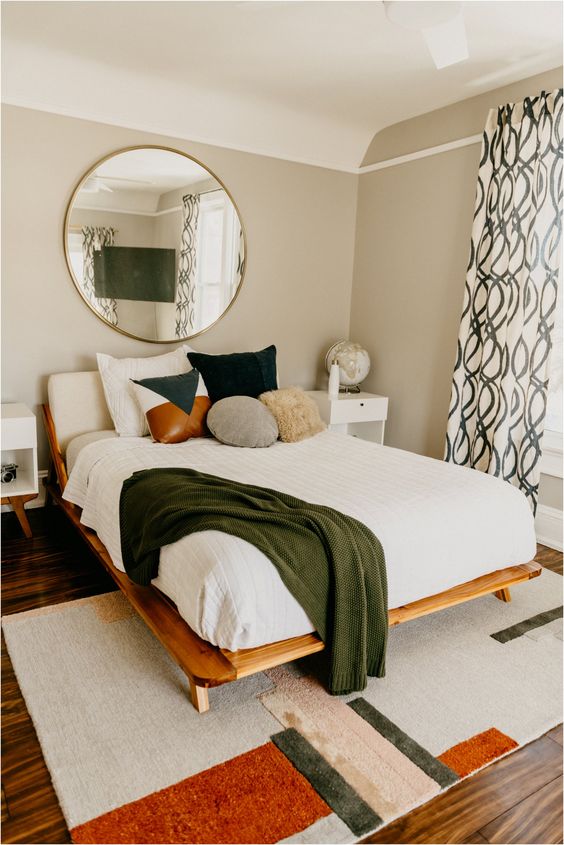 a mid-century modern bedroom with a geometric rug, a wooden bed, a round mirror, printed curtains and a variety of pillows