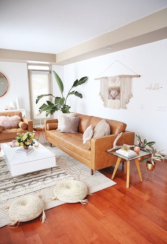 A mid century modern to boho living room with tan leather furniture, a low white coffee table, woven poufs, a macrame hanging and potted plants