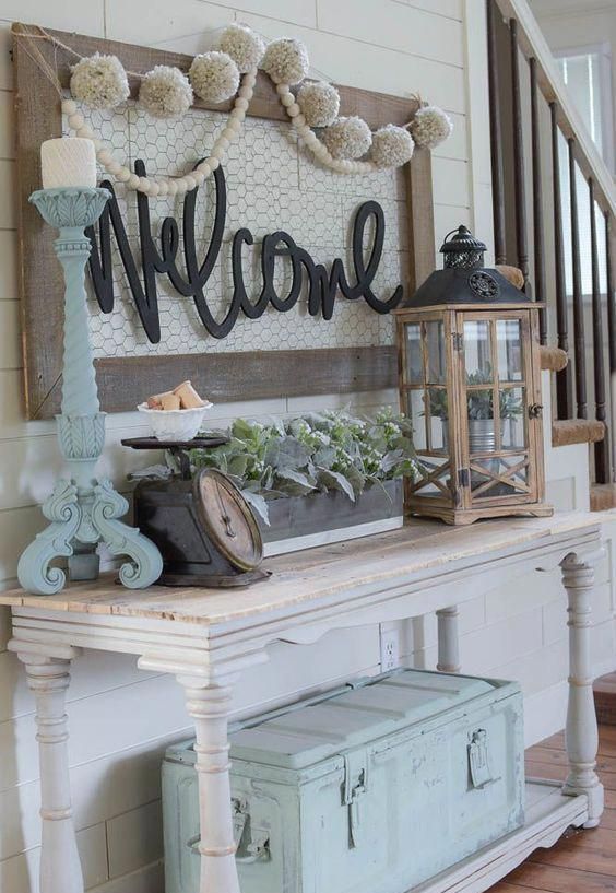 a pastel shabby chic entryway with a console table, a lantern with a pot, greenery in a box, a large chest, a candleholder and a sign with pompoms