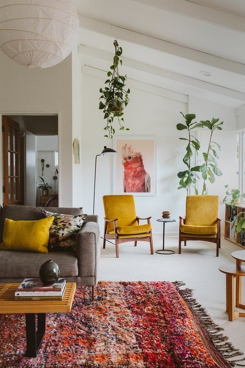 A pretty mid century modern living room with a grey sofa, mustard chairs and pillows, a colorful rug, potted plants and round tables