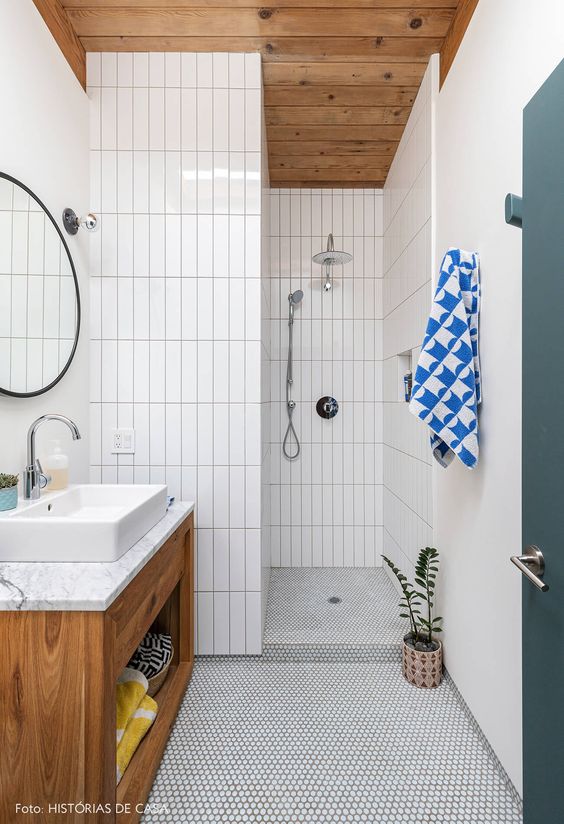 a simple mid-century modern bathroom with white penny and skinny tiles, a wooden vanity and ceiling and a round mirror