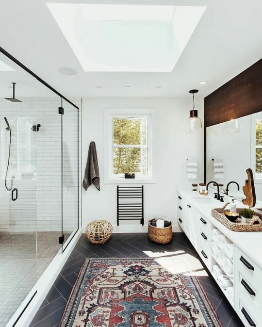 An airy mid century modern bathroom with white tiles, a skylight, a boho rug, a large vanity with a wooden top and a large shower space with windows