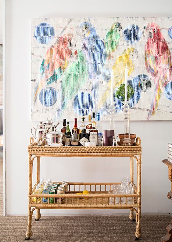 a bright space with a colorful artwork, a wicker home bar cart with lots of drinks, bottles, glasses and other stuff