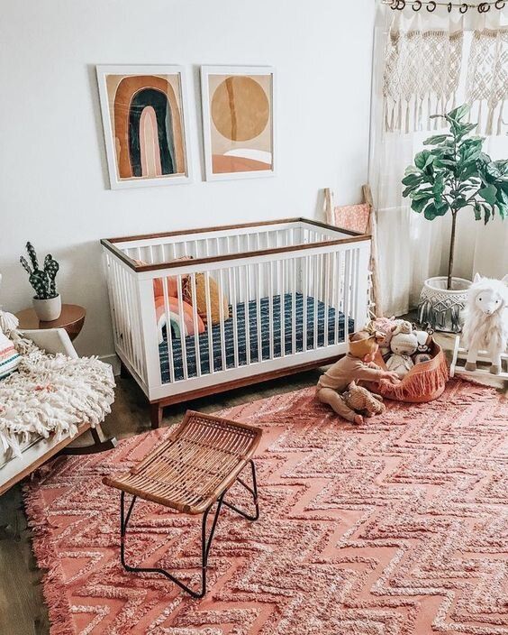 a boho girl nursery with white and rattan furniture, macrame curtains, a potted plant, a pink rug and lovely bedding