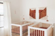 a boho twin nursery in white, with rust and orange touches, with printed bedding, a rattan chandelier and fringe artworks