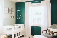 a bold modern nursery with a green accent wall, white and dark rattan furniture, layered rugs, a chandelier and a gallery wall
