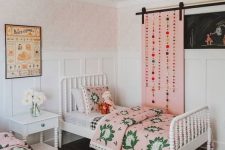 a bright shared girls’ bedroom with white vintage furniture, pink bedding and a pink barn door with pompoms, a printed rug