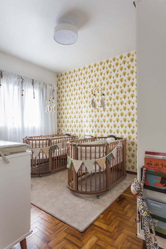 a bright shared nursery with printed wallpaper, stained cribs and neutral furniture, colorful fabric buntings and mobiles