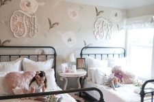 a chic farmhouse shared girls’ bedroom with a floral accent wall and monograms, with metal beds and white and pink bedding, a round table as a nightstand