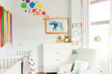 a colorful nursery with a bold wall, a colorful rug and textiles plus a wall hanging and white furniture as a backdrop