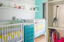 a colorful shared nursery with a grey wall, white furniture, a blue changing table, floating shelves and a colorful chandelier
