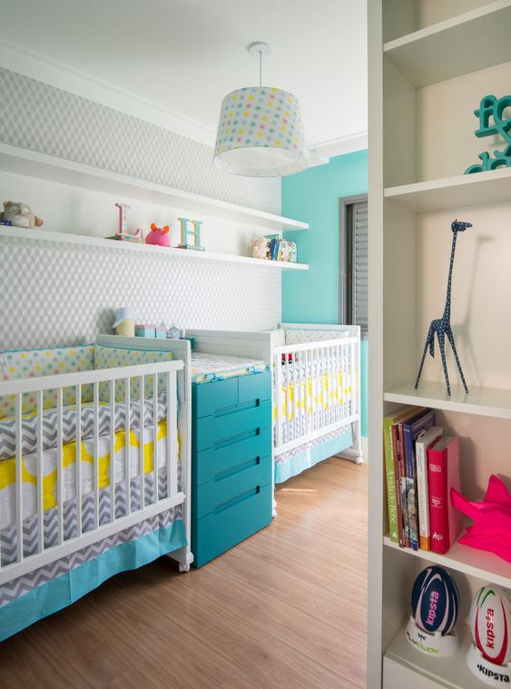 a colorful shared nursery with a grey wall, white furniture, a blue changing table, floating shelves and a colorful chandelier