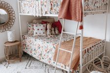 a cool boho shared girls’ bedroom with a white metal bunk bed, floral print bedding, a printed rug, a rattan chair and a nightstand, some art