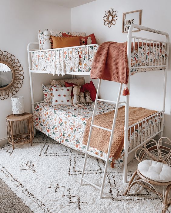 a cool boho shared girls' bedroom with a white metal bunk bed, floral print bedding, a printed rug, a rattan chair and a nightstand, some art