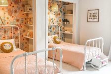 a cool shared girls’ bedroom with niches done with floral wallpaper and shelves, white metal beds, little play cribs and pink bedding