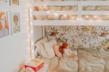 a cozy shared girls’ bedroom with a floral accent wall, a bunk bed accent with lights, a white toy chest and a gallery wall