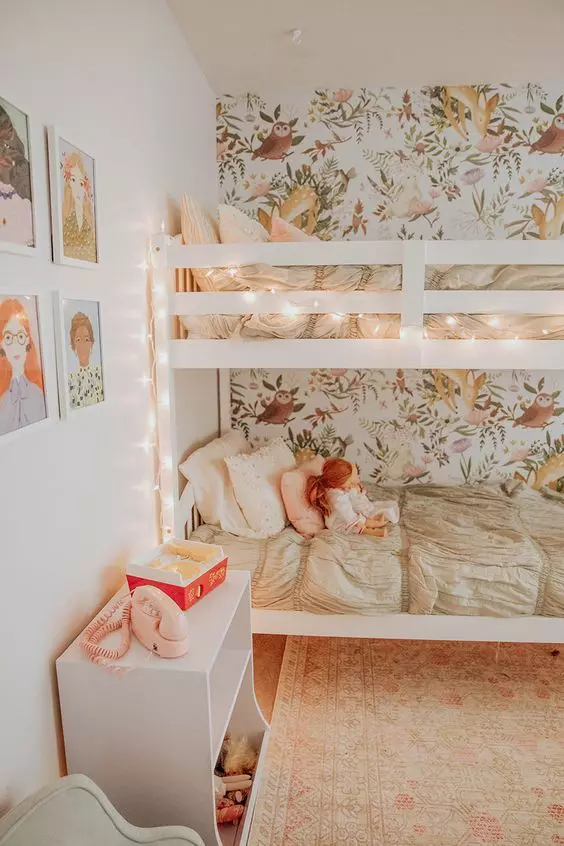 a cozy shared girls' bedroom with a floral accent wall, a bunk bed accent with lights, a white toy chest and a gallery wall
