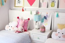 a cute and bright shared girls’ bedroom with white furniture, pastel bedding, a ledge gallery wall, a banner and colorful toys and pillows