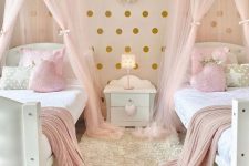 a cute and glam shared girls’ bedroom with a gold polka dot accent wall, white beds with white, pink and gold bedding, a gold rug and pink sheer canopies