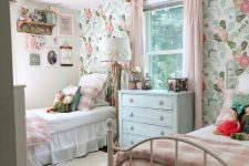 a cute shared girls’ bedroom with a pastel floral wall, white metal beds with pink and white bedding, wall-mounted shelves and toys is very cute