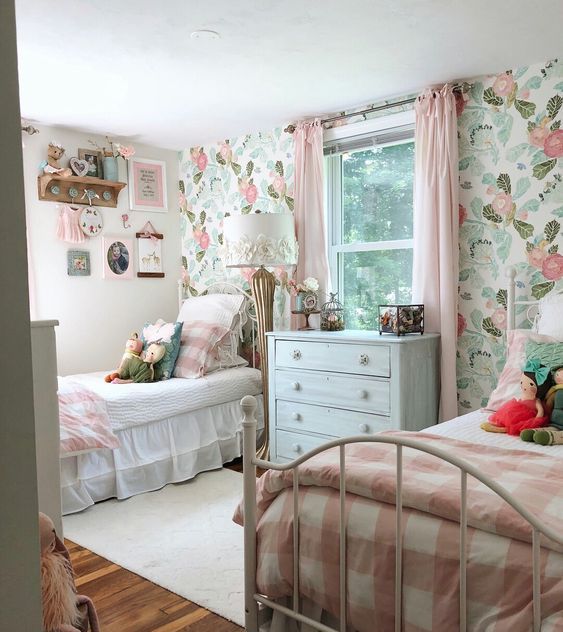 A cute shared girls' bedroom with a pastel floral wall, white metal beds with pink and white bedding, wall mounted shelves and toys is very cute