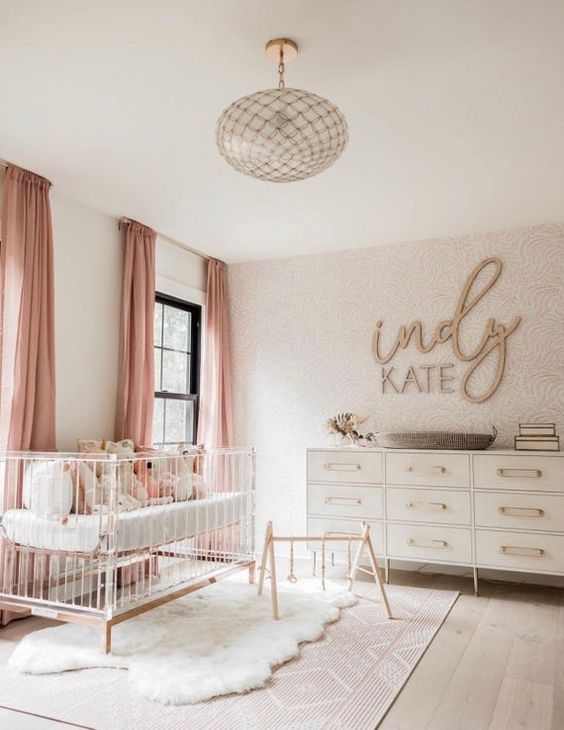 a fabulous modern girl nursery with a wallpaper wall, neutral dresser, an acryclic crib, a glass chandelier and pink textiles