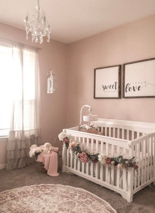 a glam girl's nursery with pink walls, white furniture, framed artworks, a floral garland and a crystal chandelier