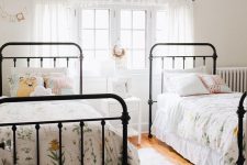 a light-filled shared girls’ bedroom with black metal beds with floral bedding, white curtains and a nightstand